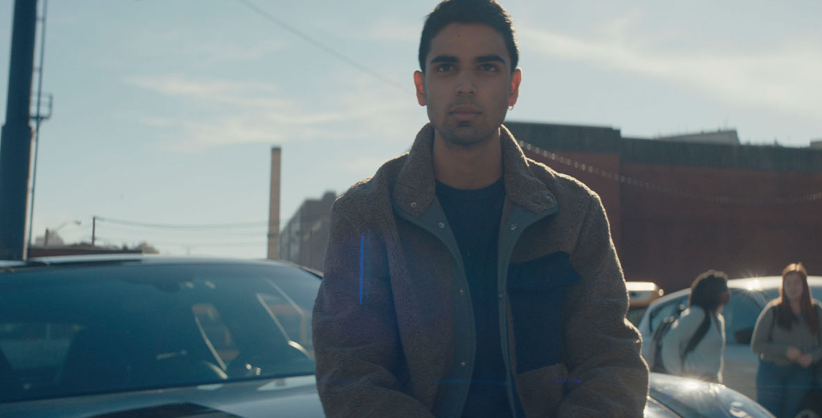 Rish Shah as Kamran, an intriguing new student who has caught Kamala’s eye, stands in front of his black Porsche in the school parking lot