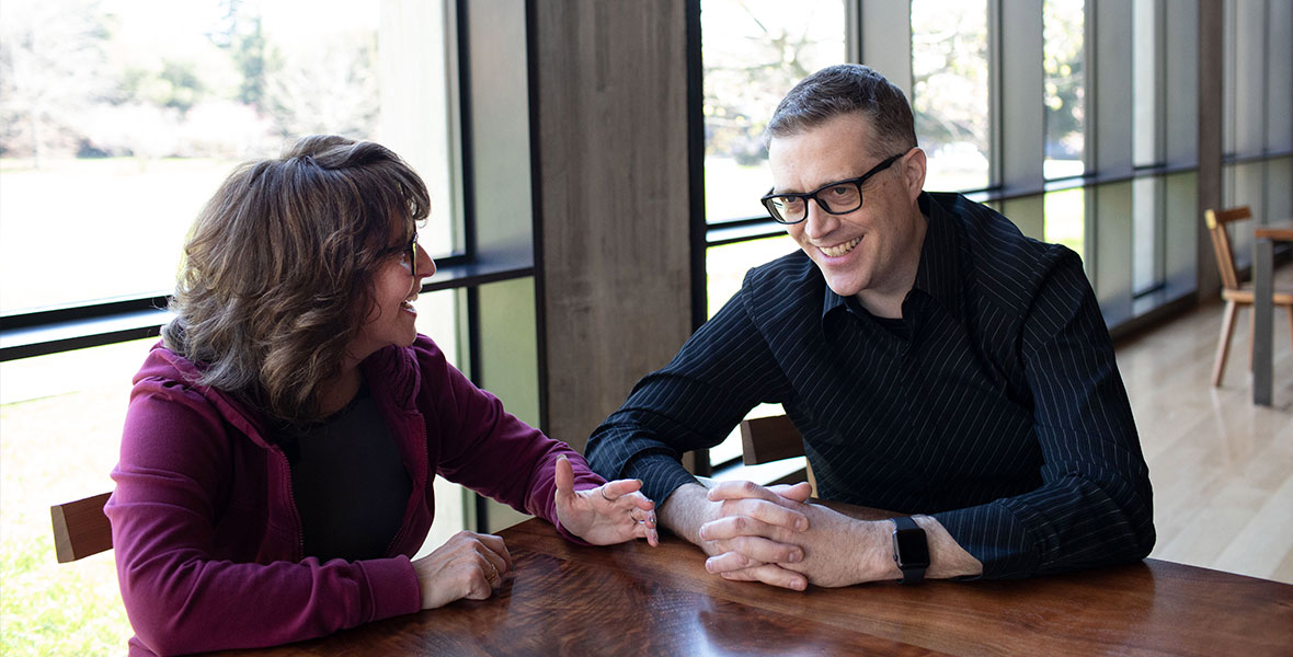 At a table, Lightyear producer Galyn Susman is wearing a purple sweater and is sitting on the left, and director Angus MacClane is wearing a dark striped button-down shirt and is sitting on the right. Galyn is looking at Angus, and Angus is looking at something off-camera; both are wearing glasses and smiling.