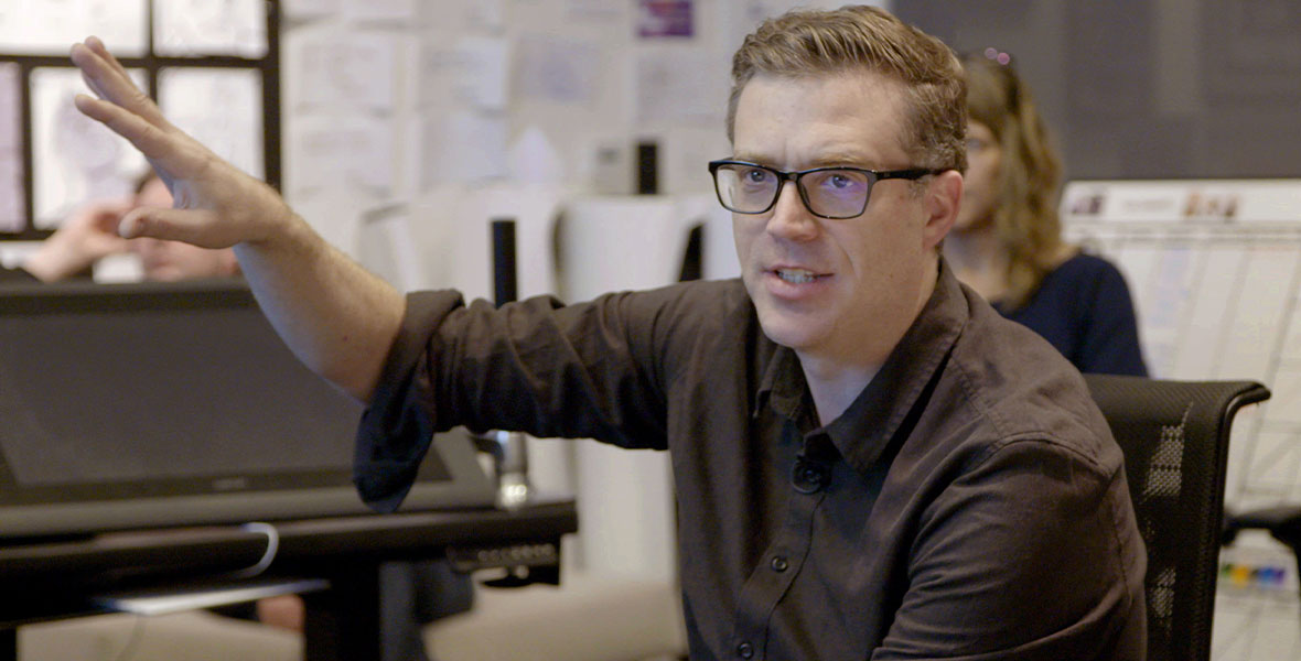 Lightyear director Angus McClane is gesturing during a notes session inside Pixar Animation Studios in Emeryville, California. There are drawings on the wall behind him, and there’s a Buzz Lightyear toy on the table to his right. He is wearing glasses and a brown button-down shirt.