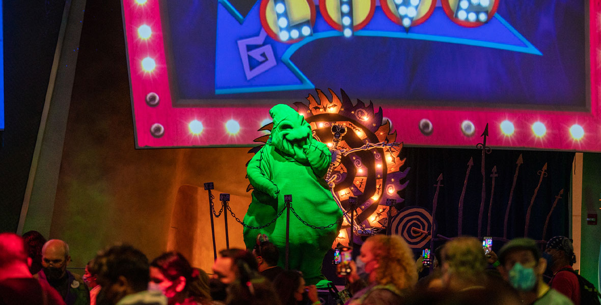 Oogie Boogie from The Nightmare Before Christmas stands above a large crowd at Disney California Adventure’s Oogie Boogie Bash. He is surrounding by neon lights and a spinning wheel with a skeleton in the middle.