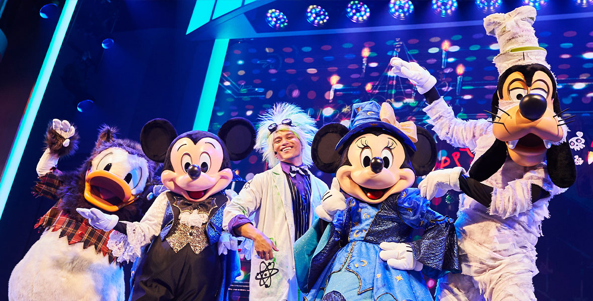 The cast of Mickey’s Trick and Treat show pose for a photo. Donald Duck is dressed as a werewolf, Mickey Mouse is dressed as a vampire, Minnie Mouse is dressed as a witch, and Goofy is dressed as a mummy. They are joined by the human host of the show, who is dressed as a mad scientist.