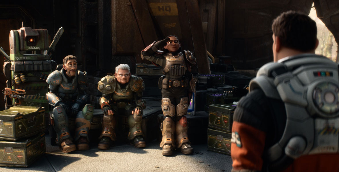 In a still from Disney and Pixar’s Lightyear, we see the back of Buzz, as he’s looking on at Izzy (who’s standing and saluting him), Darby, and Mo; all are wearing spacesuits, and they’re surrounded by boxes and other space paraphernalia.