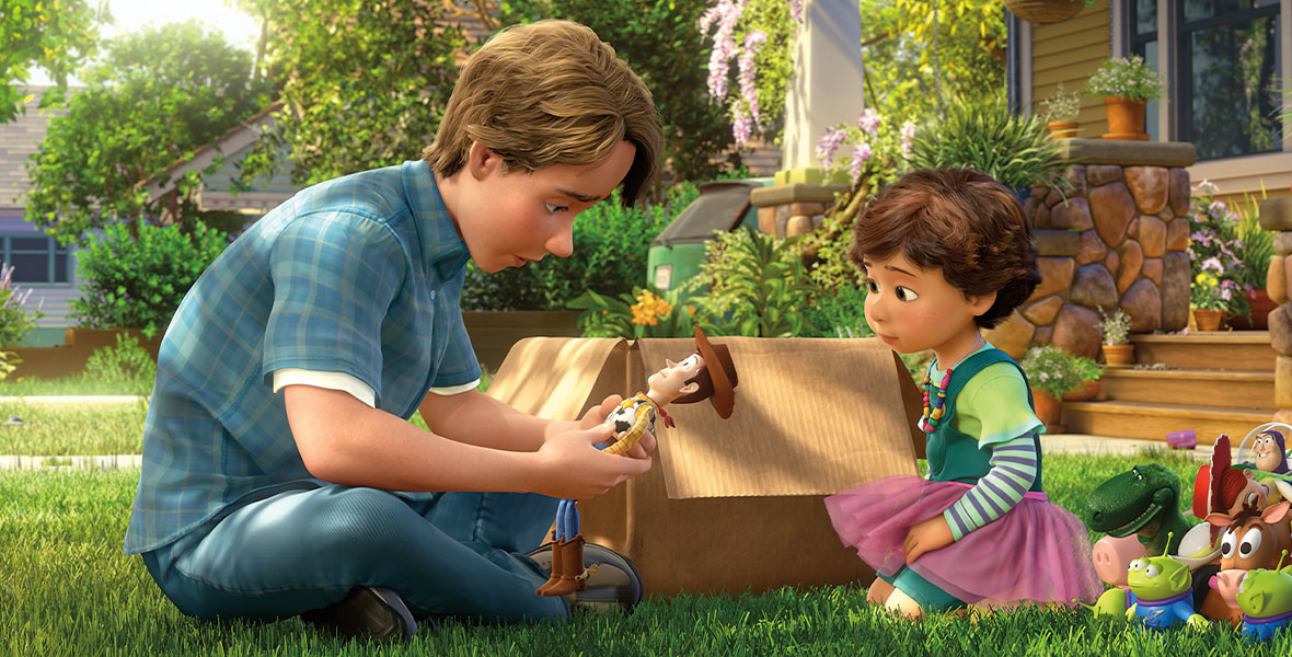 Andy (voiced by John Morris) sits on green grass looking down at his Woody and Buzz Lightyear toys whilst across from his toddler sister and an adjacent large brown box in the animated feature film Toy Story 3.