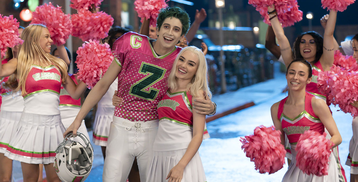 In the foreground, green-haired zombie Zed (Milo Manheim) wears a pink and green Seabrook football jersey. He has removed his helmet and is hugging blonde cheerleader Addison (Meg Donnelly), who is wearing a pink, green, and white uniform. In the background, Seabrook cheerleaders are waving pink pom poms in the air.