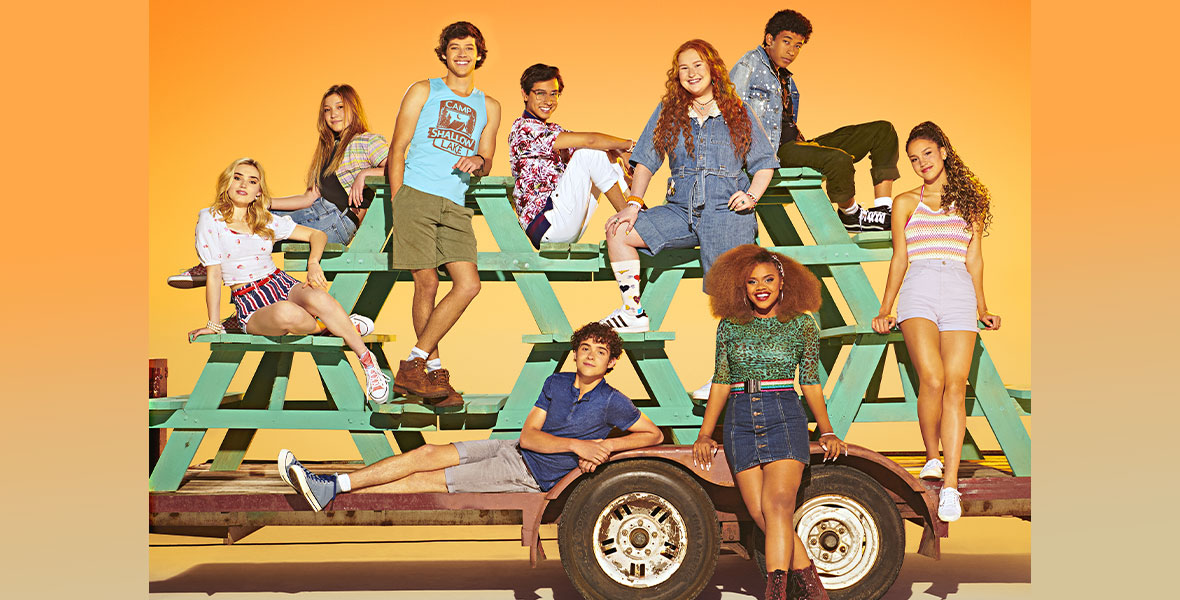 The cast of High School Musical: The Musical: The Series season three pose against an orange background. They are seated on a truck bed and stacked green picnic tables. From left to right are Meg Donnelly as Val, sitting on a picnic bench and wearing a printed white top and red, white, and blue striped shorts; Saylor Bell Curda as Maddox, sitting behind Meg on a different picnic bench and wearing a flannel shirt over a black tee and denim jeans; Matt Cornett as E.J., standing with his legs crossed and wearing a blue tank top with a brown Camp Shallow Lake logo, paired with olive green shorts and brown hiking boots; Frankie Rodriguez as Carlos, sitting on the opposite side of Saylor’s bench and wearing glasses, a red and white patterned top, and white cuffed pants; Joshua Bassett as Ricky, reclining on the truck’s wheel and wearing a blue polo, gray cuffed shorts and sneakers; Julia Lester as Ashlyn, standing behind Joshua and wearing a denim jumpsuit, patterned socks and striped sneakers; Adrian Lyles as Jet, sitting behind Julia and wearing a denim jacket, green cuffed pants and sneakers; Dara Reneé as Kourtney, sitting below Julia and wearing a green leopard print top, a rainbow striped belt, a denim skirt, and red boots; and Sofia Wylie as Gina, standing on a picnic bench and wearing a pastel crocheted top, high-waisted purple shorts and white tennis shoes. Almost everyone is smiling and looking into the camera.