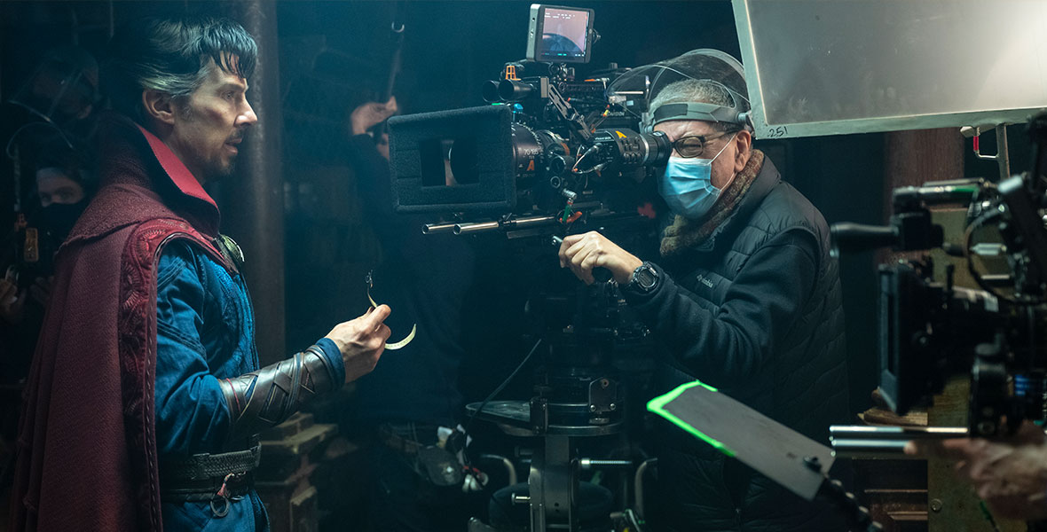 Benedict Cumberbatch wears a red cape and a blue outfit while filming a scene as Doctor Stephen Strange in Doctor Strange in the Multiverse of Madness. In front of him is A-camera operator Rodrigo Gutierrez, who is masked and squinting into the camera.