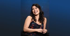 Actor Auli&rsquo;I Cravalho laughs with her arms folded in front of her, wearing a blue floral dress in front of a blue background.