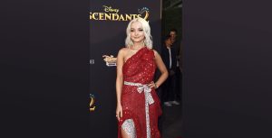Actor Dove Cameron smiles to the left of the camera on the Descendants 2 red carpet in a red dress with one hand on her hip.