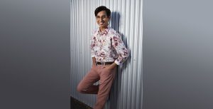 Actor Frankie Rodriguez smiles at the camera and leans against a corrugated steel wall with both hands in his pockets and one foot up on the wall.