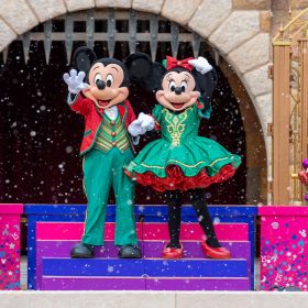 Mickey Mouse and Minnie Mouse, wearing festive red and green outfits, wave to guests from the pink, purple, and blue castle steps. They are flanked by two Christmas trees, adorned with rainbow ornaments and topped with gold and white stars, as well as two snowman pals, wearing scarves, hats, and boots to keep warm amid the snowfall.