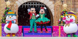 Mickey Mouse and Minnie Mouse, wearing festive red and green outfits, wave to guests from the pink, purple, and blue castle steps. They are flanked by two Christmas trees, adorned with rainbow ornaments and topped with gold and white stars, as well as two snowman pals, wearing scarves, hats, and boots to keep warm amid the snowfall.