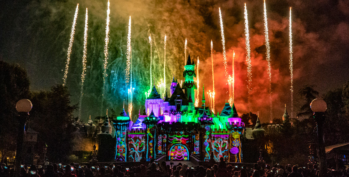 Fireworks light up the sky in rainbow colors in front of Cinderella Castle at Disneyland park. Images of neon gumdrops, peppermints, candy canes, and lollipops are projected onto the castle. In the foreground, guests marvel at the “Believe.... in Holiday Magic” fireworks spectacular.