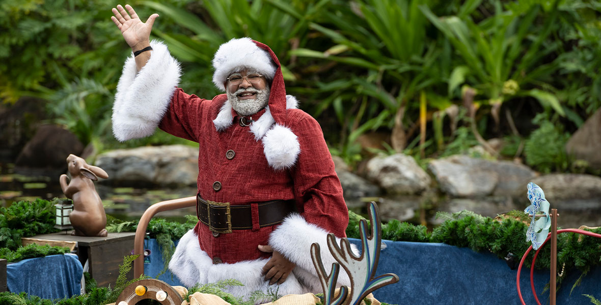 Santa Claus smiles wide and wears his signature red outfit, featuring white fur trim. He is standing a parade float and waving to guests at Walt Disney World Resort.