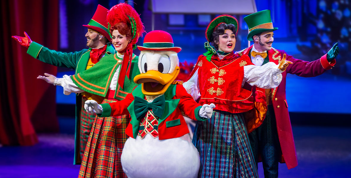 Donald Duck, wearing a red hat and jacket with green trim and a large green bowtie, stands centerstage during the musical show “Let’s Sing Christmas!” at Videopolis in Discoveryland at Disneyland Paris. Donald is flanked by four carolers, each wearing festive, old-timey red and green attire, as they smile at the audience.