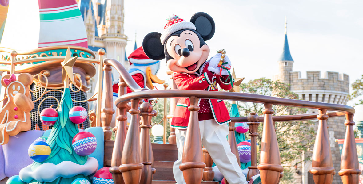 Mickey Mouse stands on a parade float. He’s wearing a blazer with green and white trim, a red and white knit hat, and white pants. He’s holding bells and smiling.