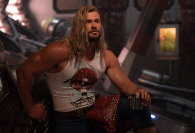 In a production still from Marvel Studios’ Thor: Love and Thunder, Thor (Chris Hemsworth) relaxes inside the Guardians of the Galaxy’s starship, the Milano. He is flexing his biceps and holding a cup, with his leg propped up. His hair is long and features a single braid, and he wears a tight white tank top with dark pants. His studded red leather jacket is draped over his leg.