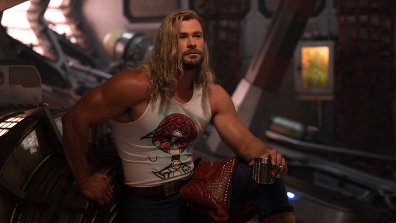 In a production still from Marvel Studios’ Thor: Love and Thunder, Thor (Chris Hemsworth) relaxes inside the Guardians of the Galaxy’s starship, the Milano. He is flexing his biceps and holding a cup, with his leg propped up. His hair is long and features a single braid, and he wears a tight white tank top with dark pants. His studded red leather jacket is draped over his leg.