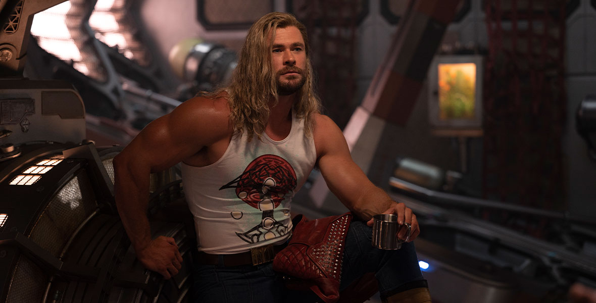 In a production still from Marvel Studios’ Thor: Love and Thunder, Thor (Chris Hemsworth) relaxes inside the Guardians of the Galaxy’s starship, the Milano. He is flexing his biceps and holding a cup, with his leg propped up. His hair is long and features a single braid, and he wears a tight white tank top with dark pants. His studded red leather jacket is draped over his leg.