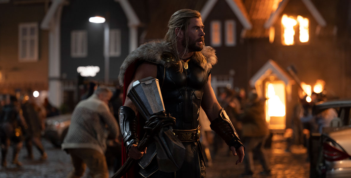 Alt Text: In a production still from Marvel Studios’ Thor: Love and Thunder, Thor (Chris Hemsworth) is wearing his classic Asgardian battle armor, including a fur-trimmed red cape and leather arm cuffs. He is holding Stormbreaker, his magical axe, and gazing into the distance. Behind him, the citizens of New Asgard are under attack.