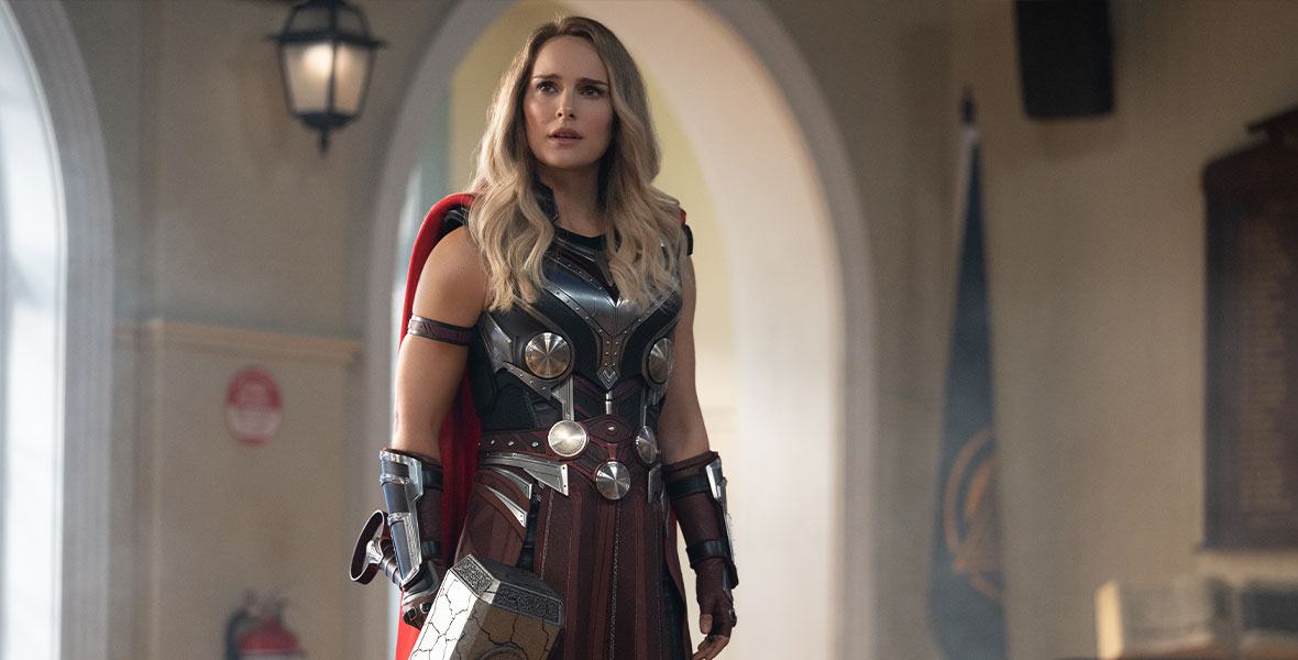Alt Text: In a production still from Marvel Studios’ Thor: Love and Thunder, The Mighty Thor (Natalie Portman) stands tall in full Asgardian regalia. Now blonde, she wears a flowing red cape, a leather skirt, and a metallic armor. She holds Mjolnir, Thor’s magical hammer, in her right hand.