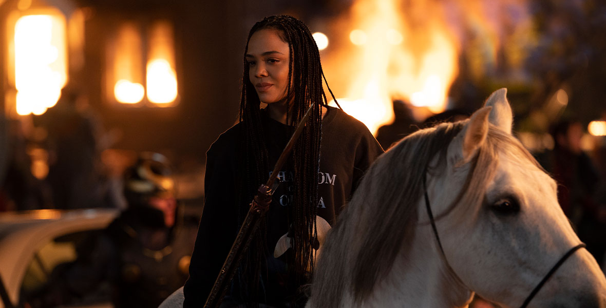 In a production still from Marvel Studios’ Thor: Love and Thunder, King Valkyrie (Tessa Thompson) is on horseback as New Asgard is under attack. She is wearing a Phantom of the Opera sweatshirt and is holding a spear.