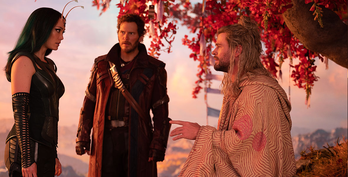 In a production still from Marvel Studios’ Thor: Love and Thunder, Mantis (Pom Klementieff), Star-Lord (Chris Pratt), and Thor (Chris Hemsworth) are having an intense conversation. Mantis is standing opposite Thor, who is sitting and meditating under a tree; Star-Lord stands between them and is looking at Mantis. Mantis is wearing an all-black leather outfit. Star-Lord is wearing a red leather trench coat. Thor is wearing a patterned tunic and his messy blonde hair is partially pulled into a bun.