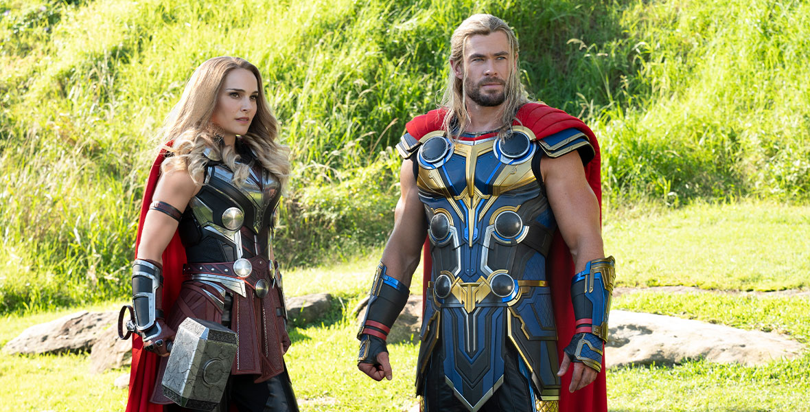 In a production still from Marvel Studios’ Thor: Love and Thunder, The Mighty Thor (Natalie Portman) stands to the left of Thor (Chris Hemsworth) in a grassy field. They are wearing variations of the classic Thor costume, which includes Asgardian battle armor and red capes, and they both have long blonde hair. The Mighty Thor wields the hammer Mjolnir, which shows fracture lines from where it had been broken.