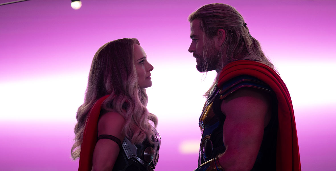 In a production still from Marvel Studios’ Thor: Love and Thunder, The Mighty Thor (Natalie Portman) stands to the left of Thor (Chris Hemsworth) against a purple background. They are wearing variations of the classic Thor costume, which includes Asgardian battle armor and red capes, and they both have long blonde hair. Their arms are muscular and toned, and they are gazing into the other person’s eyes.