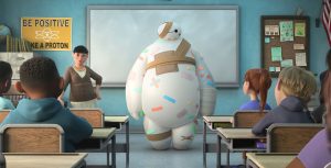 Baymax, the inflatable healthcare companion robot, is covered in multicolored bandages. He is standing at the front of a classroom next to a teacher. Behind them is a whiteboard and a poster that reads: “Be Positive Like a Proton.” In the foreground, students sit at their desks, with their attention turned towards Baymax.