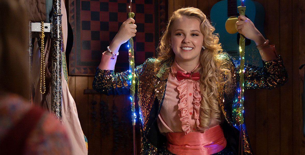 JoJo Siwa guest stars in the third season of High School Musical: The Musical: The Series. In this scene, she wears a sequined suit with a pink dress shirt, a pink cummerbund, and a pink bowtie. She is sitting on a swing illuminated by string lights.