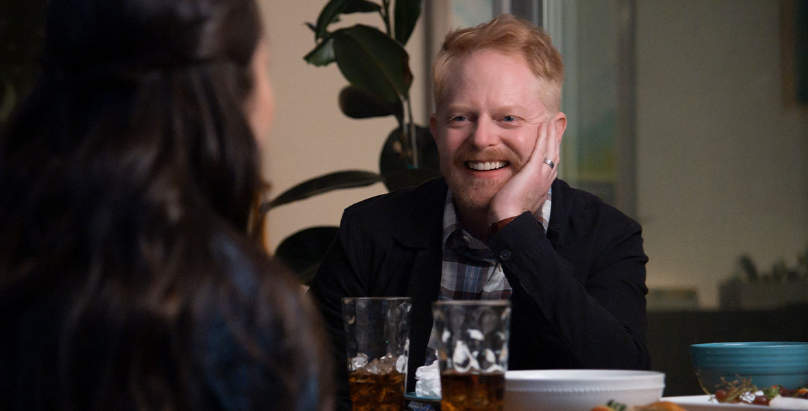 Jesse Tyler Ferguson guest stars in the third season of High School Musical: The Musical: The Series. In this scene, he is wearing a black sweater over a plaid shirt. He is resting his chin in his hand and seated at a dinner table across from Olivia Rodrigo as Nini, whose back is to the camera.