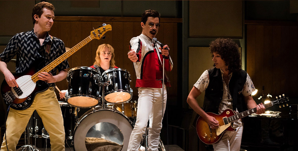 Freddie Mercury, who is wearing an all-white suit, stands at center singing with his band Queen in a recording studio in the film Bohemian Rhapsody.