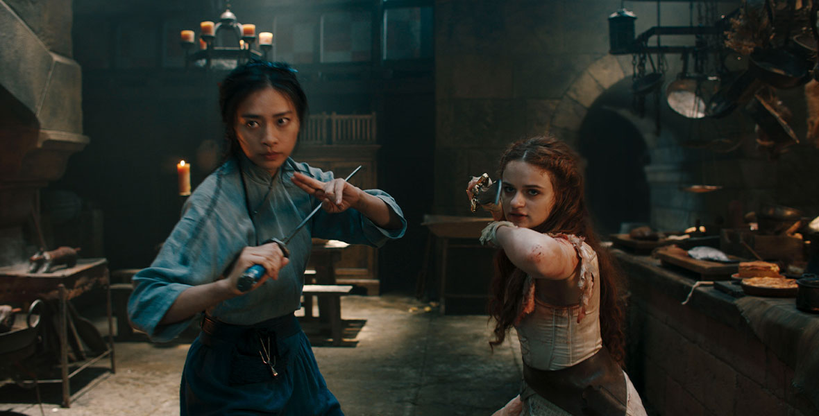 Veronica Ngo and Joey King stand with swords in the castle’s kitchen in a fighting stance.