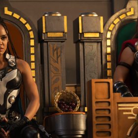 A still from Thor: Love & Thunder featuring Valkyrie, portrayed by actress Tessa Thompson, and Jane Foster, portrayed by actress Natalie Portman, site next to each other on ornate thrones with gold accents. Both wear armor and serious expressions.