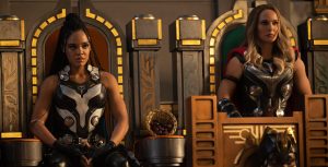 A still from Thor: Love & Thunder featuring Valkyrie, portrayed by actress Tessa Thompson, and Jane Foster, portrayed by actress Natalie Portman, site next to each other on ornate thrones with gold accents. Both wear armor and serious expressions.