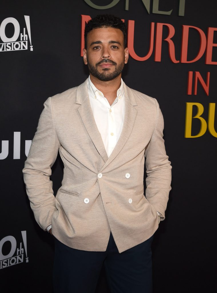Ryan Broussard on the red carpet at the premiere of Hulu’s Only Murders in the Building season 2. He’s wearing a tan suit jacket and black pants. The logo for the show, as well as the 20th Century Television and Hulu logos, are seen behind him.