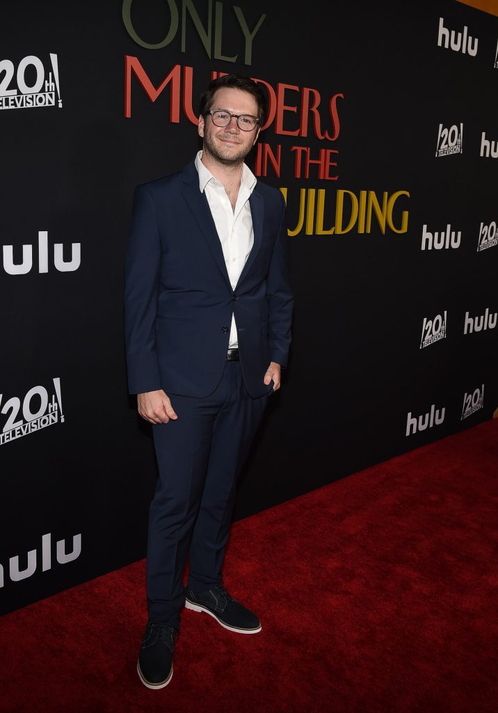 James Caverly on the red carpet at the premiere of Hulu’s Only Murders in the Building season 2. He’s wearing a dark blue suit and glasses. The logo for the show, as well as the 20th Century Television and Hulu logos, are seen behind him.