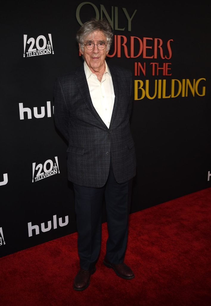 Elliott Gould on the red carpet at the premiere of Hulu’s Only Murders in the Building season 2. He is wearing a dark suit and glasses. The logo for the show, as well as the 20th Century Television and Hulu logos, are seen behind him.