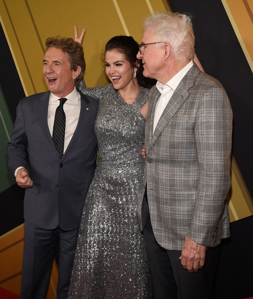 Left to right: Martin Short (in a dark grey suit), Selena Gomez (in a sparkly silver gown), and Disney Legend Steve Martin (in a plaid suit jacket) laughing together on the red carpet at the premiere of Hulu’s Only Murders in the Building season 2. Gomez is making “bunny ears” with her fingers behind Short’s head.
