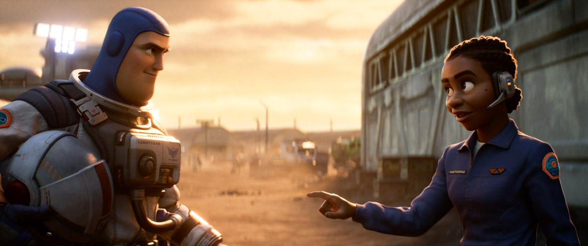 Buzz (wearing a spacesuit and holding a helmet) is standing on the left, while Alisha (wearing a blue jumpsuit and a headset) is pointing her index finger towards Buzz and is standing on the right, in a still from Disney and Pixar’s Lightyear. There are several buildings and large floodlights in the background.