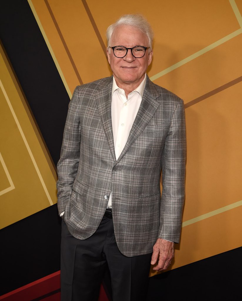 Disney Legend Steve Martin on the red carpet at the premiere of Hulu’s Only Murders in the Building season 2. He’s wearing a grey plaid suit jacket and glasses and standing in front of a yellow and black background.