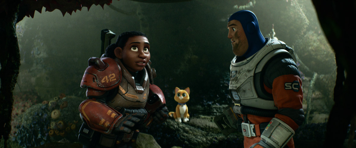 Izzy is wearing a spacesuit and is looking up at something off-camera, standing to the left of Buzz, who’s also wearing a spacesuit and is looking at Izzy with some concern, in a still from Disney and Pixar’s Lightyear. Buzz has dirt or oil of some kind on his face. Sox the cat robot companion can be seen in the background between them. They appear to be in a forest.