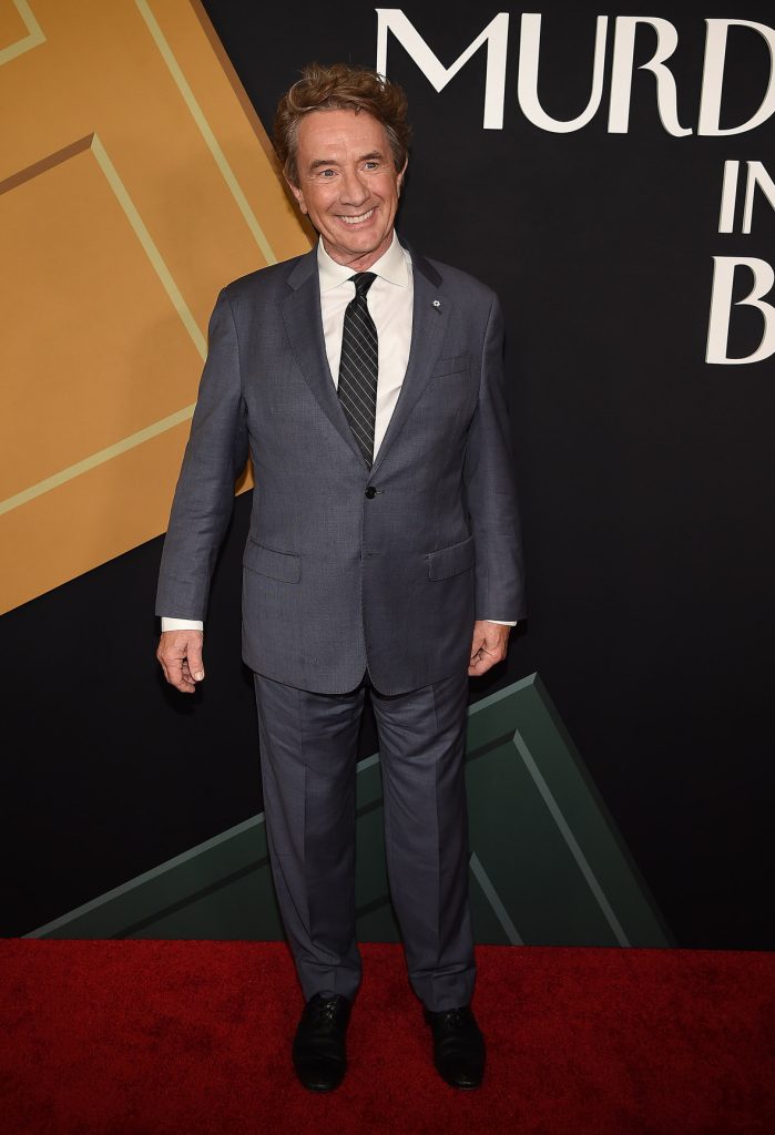Martin Short on the red carpet at the premiere of Hulu’s Only Murders in the Building season 2. He’s wearing a dark grey suit with a grey striped tie, and is standing in front of a background that includes the show’s logo.