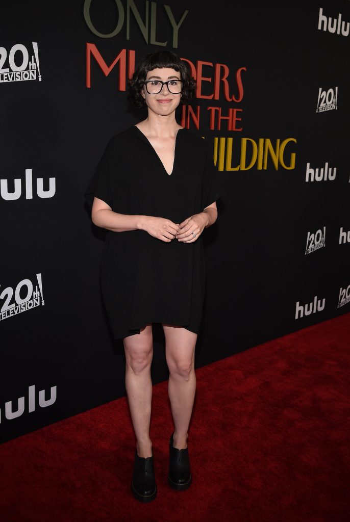 Adina Verson on the red carpet at the premiere of Hulu’s Only Murders in the Building season 2. She’s wearing a black dress and glasses. The logo for the show, as well as the 20th Century Television and Hulu logos, are seen behind her.