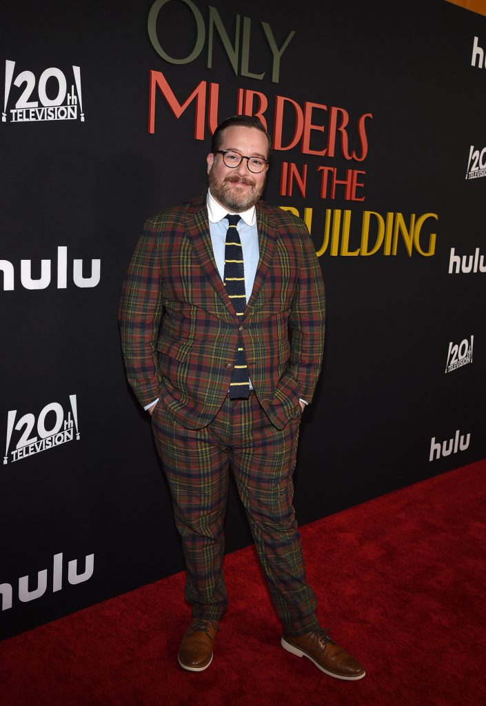 Michael Cyril Creighton on the red carpet at the premiere of Hulu’s Only Murders in the Building season 2. He’s wearing a brown tweed suit and glasses. The logo for the show, as well as the 20th Century Television and Hulu logos, are seen behind him.