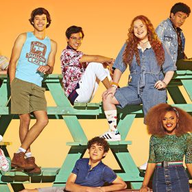 The cast of Disney+’s High School Musical: The Musical: The Series season three is seen sitting and standing on green-painted picnic tables that are piled on the back of a flatbed with large wheels, up against an orange background. Each actor is wearing summery clothing, like jeans, shorts, and T-shirts.