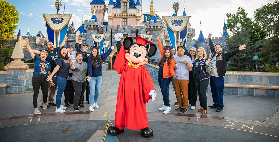 Mickey Mouse, wearing a red graduation gown and cap and a yellow bowtie, pumps his right fist in the air. He is standing in front of Cinderella Castle at Magic Kingdom at Walt Disney World Resort. Behind him stand 13 employees who are enrolled in the Disney Aspire program; their arms are outstretched and they are smiling. The employees are wearing jeans and shirts that represent their alma matters.