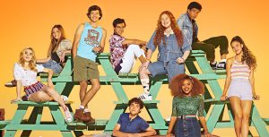 The cast of High School Musical: The Musical: The Series Season 3 sitting on top of picnic tables that are stacked on top of a trailer bed. There are about five stacked picnic tables and the cast is sitting and standing all around. From left to right on the top row: Meg Donnelly, blonde, sitting casually in a white top and red, white, and blue striped shorts with white and red sneakers; Saylor Bell, long auburn straight hair, wearing jeans, a black tank top, and a colorful short sleeved cardigan; Matt Cornett, brunette, standing in green cargo shorts, brown work boots, and a blue tank top that says “Camp Shallow Lake” on it’ Frankie Rodriguez sitting in white ¾ length pants and a red and white patterned button up shirt. He has glasses on and has short brown hair; Julia Lester standing with on leg up on a ledge. She has long, curly red hair and is wearing a denim jumpsuit. She has on lots of bangle bracelets and has heart-patterned socks on with black and white Adidas; Adrian Lyles sitting and wearing a denim jacket, green pants, and black sneakers. He has short, black curly hair. On the bottom row: Joshua Basset laying down with his torso propped up on the wheel of the tractor. He is wearing a blue polo shirt, gray jean shorts, and like blue Converse-style shoes. He has curly, brown hair; Dara Reneé standing in a jean skirt and green leopard print shirt. She has voluminous red hair and is wearing silver hoop earrings and sparkly red boots; Sofia Wylie standing against a picnic table in lavender shorts and a crochet knit striped tank top. She has braided hair in a ponytail that ends is loose curls.