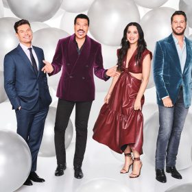 In a promotional still for ABC’s American Idol, the judges and host are standing against a backdrop of silver and white balloons. From left to right: Ryan Seacrest is wearing a blue suit; Lionel Richie is wearing a purple velvet blazer and dark pants; Katy Perry is wearing a shiny burnt-orange dress; and Luke Bryan is wearing a blue velvet blazer and gray jeans.