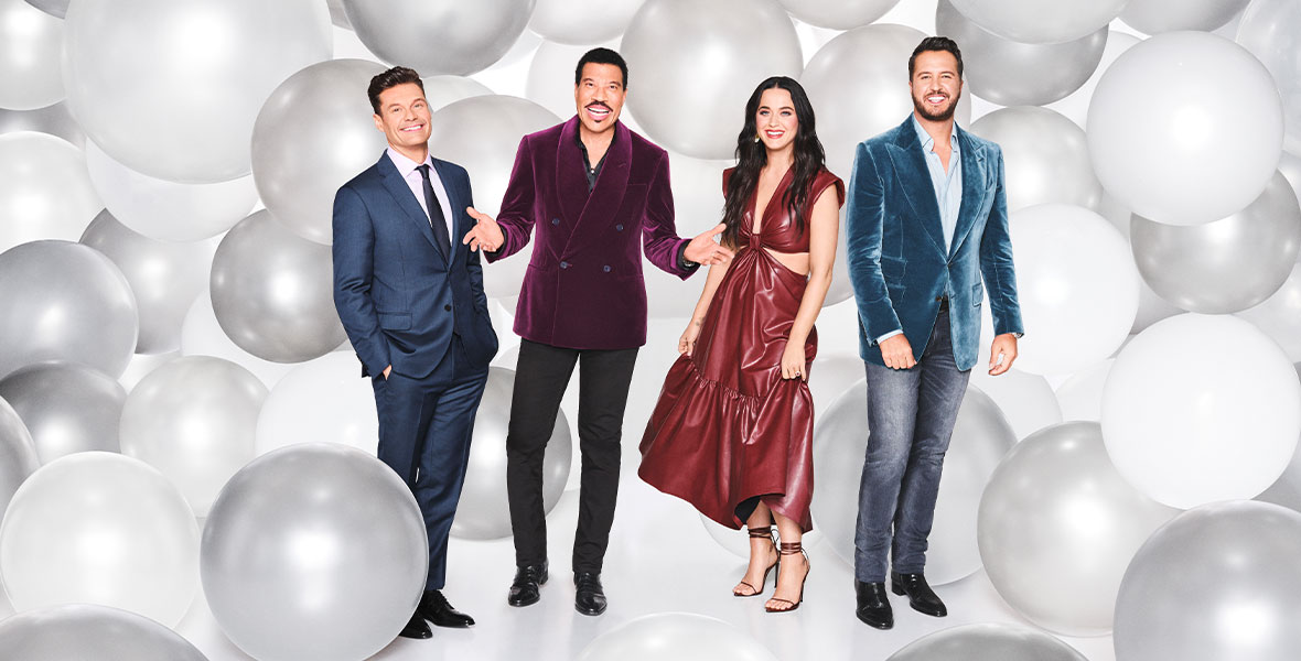 In a promotional still for ABC’s American Idol, the judges and host are standing against a backdrop of silver and white balloons. From left to right: Ryan Seacrest is wearing a blue suit; Lionel Richie is wearing a purple velvet blazer and dark pants; Katy Perry is wearing a shiny burnt-orange dress; and Luke Bryan is wearing a blue velvet blazer and gray jeans.
